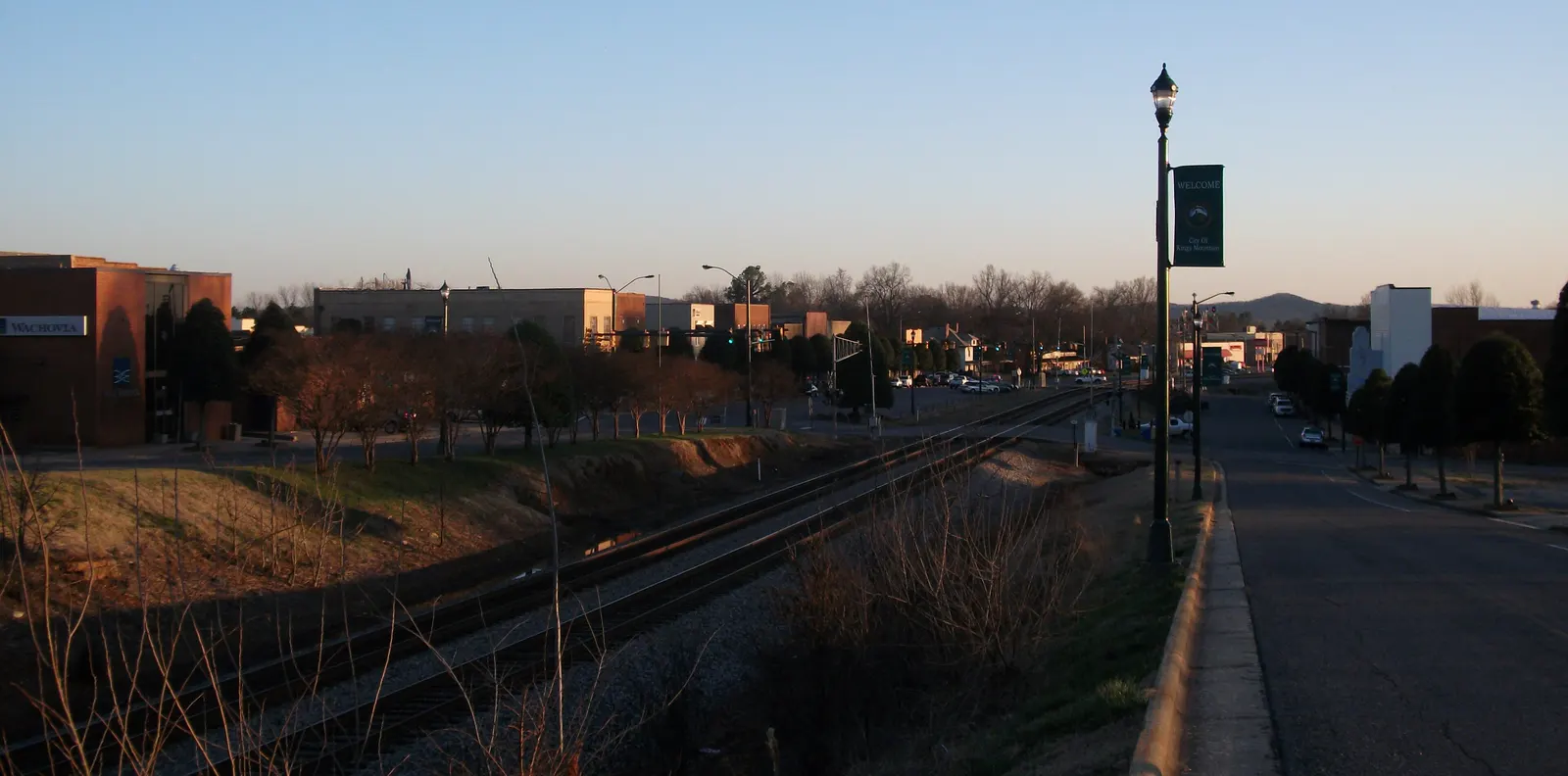Downtown Kings Mountain at dusk: brick commercial buildings, a lamp post with a city welcome banner and the railroad running past